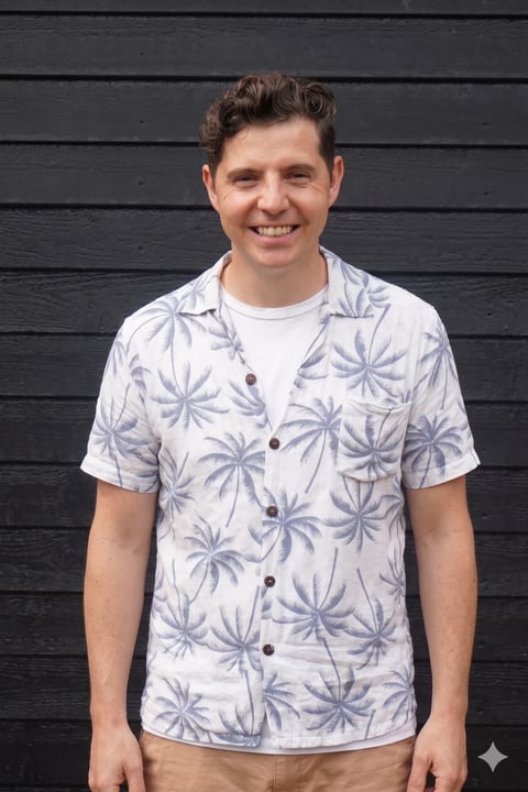 Young man smiling at camera wearing a white palm tree print shirt against a black wooden wall background